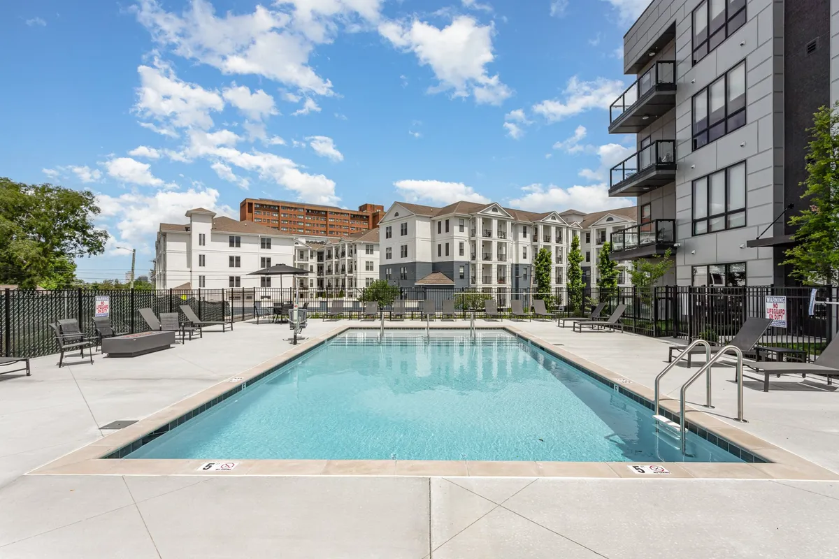 This image showcases a sparkling swimming pool surrounded by sun loungers and modern apartment buildings, with lush landscaping providing a relaxed, resort-like atmosphere. The setting suggests both opportunities for swimming and socializing by the poolside.
