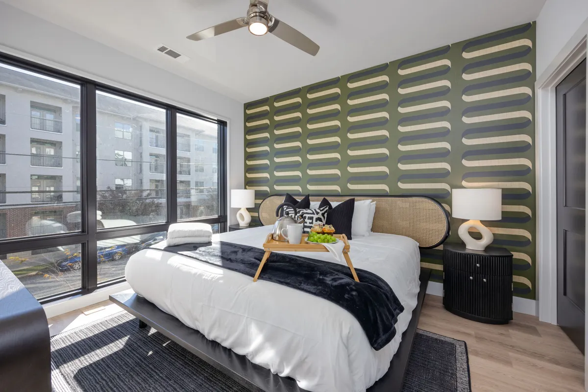 A cozy bedroom with a decorative throw on the bed, a green patterned accent wall, and large windows letting in streams of natural light. Side tables with lamps flank the bed, creating a peaceful and stylish sleeping environment.
