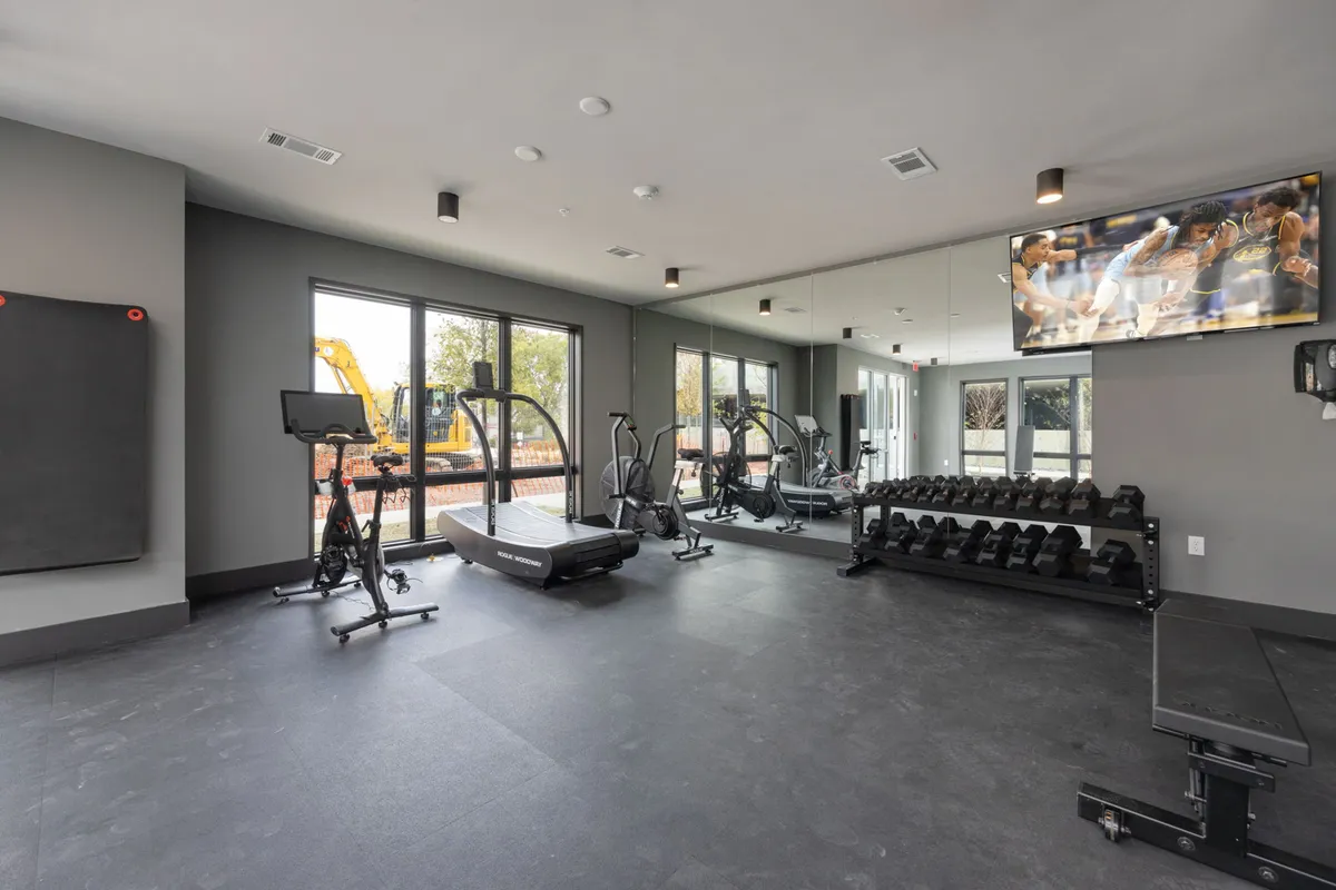A bright, well-equipped fitness center featuring a treadmill, exercise bike, free weights, large windows, and a wall-mounted TV. The room exudes energy and provides a glimpse into healthy routines available on-site.