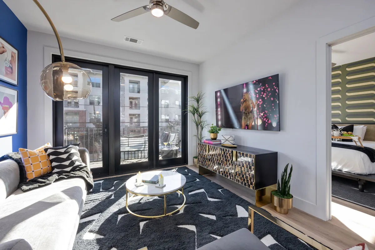 A spacious living area featuring a comfortable sectional sofa adorned with patterned throw pillows, a round coffee table, large windows offering a view of the city, and a wall-mounted television with a vibrant display. The space is bright and airy, thanks to the abundance of natural light streaming through the floor-to-ceiling windows.