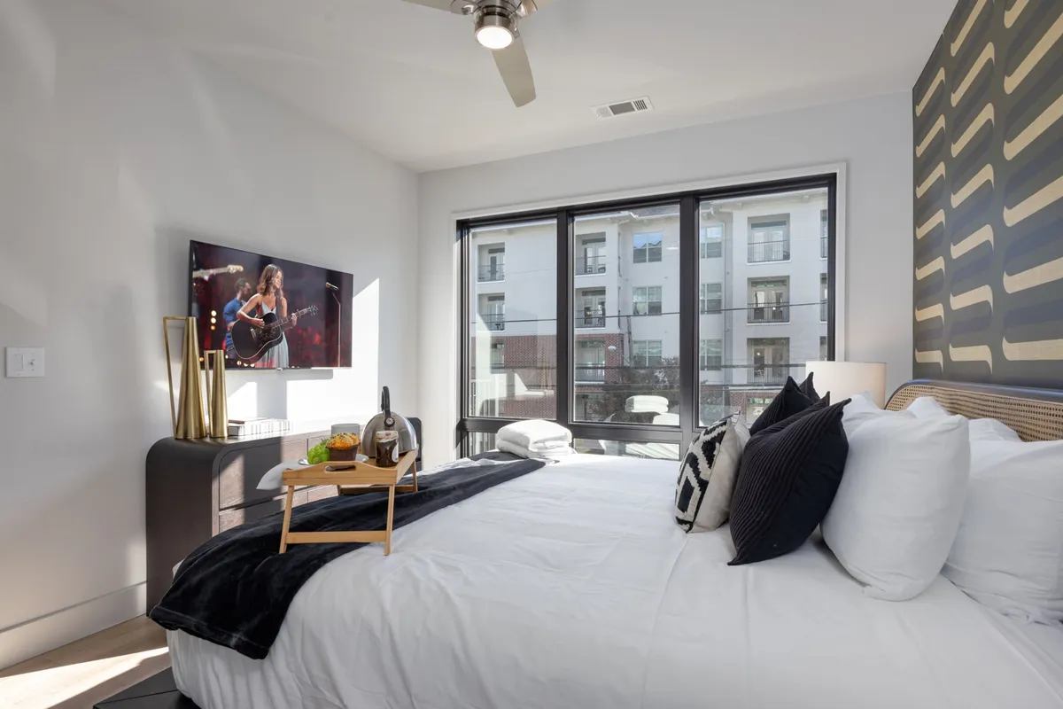 A bright, airy bedroom featuring a plush king bed with crisp white linens, a large wall-mounted television displaying a performance, decorative pillows, a sleek black accent wall, and large windows letting in abundant natural light.