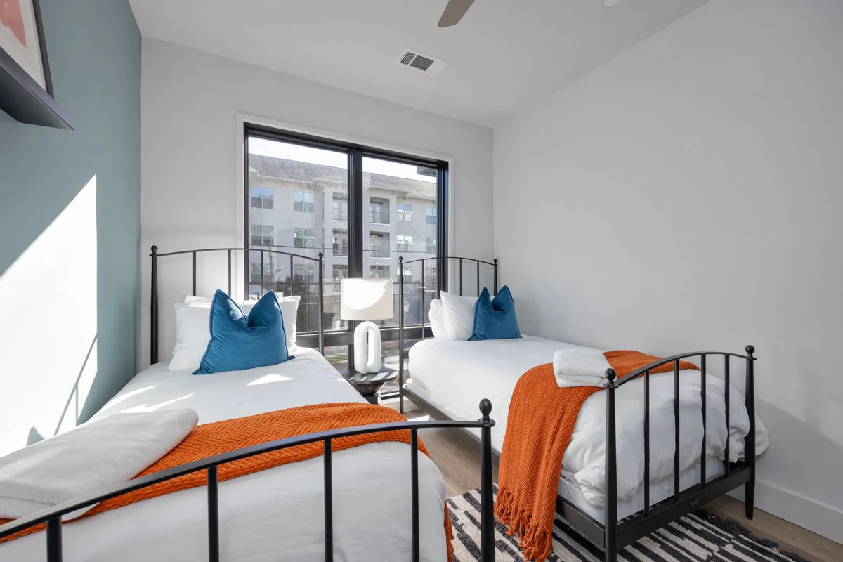 A cheerful second bedroom with two twin beds dressed in vibrant orange throws and blue pillows, large windows streaming in natural light, and a relaxing, friendly ambiance.